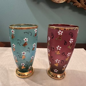 K&ouml;nnte beinhalten: Zwei Glasvasen mit goldenen R&auml;ndern und Blumenmustern. Die Vase links ist hellblau mit wei&szlig;en Blumen und goldenen Akzenten. Die Vase rechts ist burgunderrot mit wei&szlig;en Blumen und goldenen Akzenten.