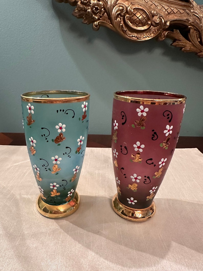 K&ouml;nnte beinhalten: Zwei Glasvasen mit goldenen R&auml;ndern und Blumenmustern. Die Vase links ist hellblau mit wei&szlig;en Blumen und goldenen Akzenten. Die Vase rechts ist burgunderrot mit wei&szlig;en Blumen und goldenen Akzenten.
