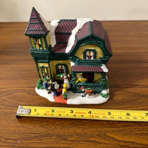 May include: A ceramic Christmas village house with a green and yellow exterior, a red roof with white snow, and a porch with figures of people. The house is 4 inches tall.