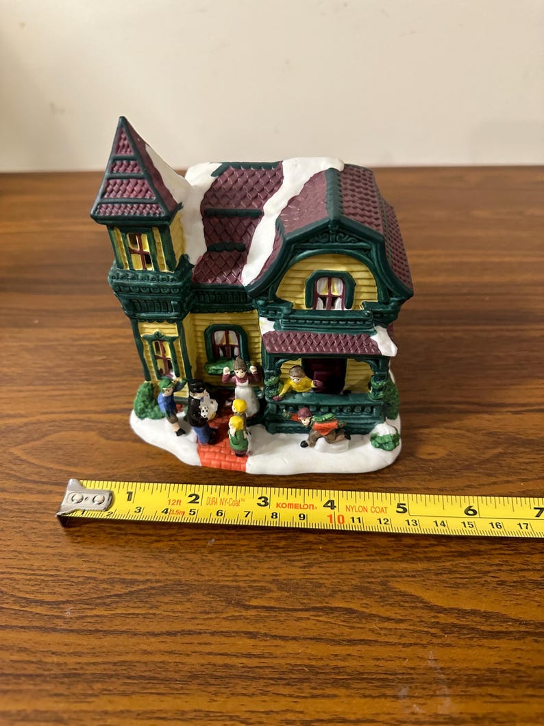 May include: A ceramic Christmas village house with a green and yellow exterior, a red roof with white snow, and a porch with figures of people. The house is 4 inches tall.