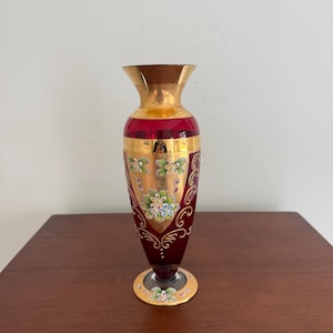 May include: A ruby red glass vase with gold accents and floral designs. The vase has a flared gold rim and a gold base. The floral designs are in shades of blue, green, and white. The vase is approximately 20 cm tall.