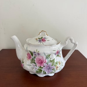 May include: A white porcelain teapot featuring a scalloped rim and a matching lid with a gold-trimmed handle. The body and lid are decorated with a colorful floral pattern consisting of pink roses, purple flowers, and green leaves on a white base.