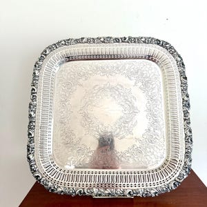 Pode incluir: Uma bandeja retangular prateada com detalhes ornamentados de flores e volutas. A bandeja tem uma borda decorativa com um design perfurado e uma borda elevada. A bandeja é provavelmente feita de metal e é adequada para servir alimentos ou exibir itens.