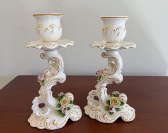 May include: A pair of white ceramic candle holders with gold trim and floral accents. The candle holders have an ornate design with a base of small flowers and leaves. The candle holders are approximately 15 cm tall.