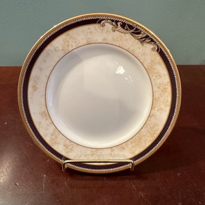 Puede incluir: Un plato de porcelana blanca con un borde dorado y negro. El plato tiene un diseño decorativo de remolinos dorados y un borde de cuerda.