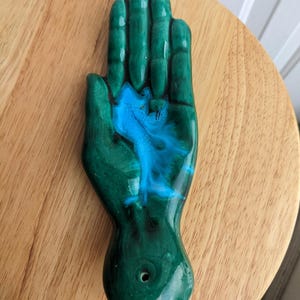 Puede incluir: Una pieza de cerámica con forma de mano en tonos verdes y azules. La zona de la palma presenta un diseño azul vibrante, mientras que los dedos y la base son de un verde brillante. Se ve un pequeño agujero en la base. La pieza está colocada sobre una superficie de madera.