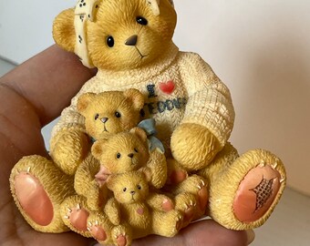May include: A figurine of a mother bear and three cubs. The mother bear wears a sweater with "I Teddy" on it and a bow. The bears are light brown with pink accents on their paws. The figurine is held in a hand.