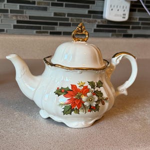 Peut inclure: Une théière en céramique couleur crème ornée d'un motif floral festif de poinsettias et de baies de houx. La théière présente un corps festonné, un bord doré et un bouton décoratif doré sur le couvercle, avec une anse et un bec courbés.