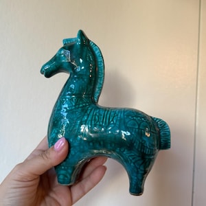 Puede incluir: Una figura de caballo de cerámica color verde azulado con un acabado brillante y complejos patrones grabados en tonos oscuros que cubren su cuerpo. El caballo se muestra en posición de pie con una crin y cola estilizadas, de color intenso.