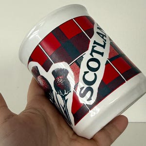 May include: White ceramic mug with a red and black tartan pattern and the word "SCOTLAND" in white. The mug features a thistle design. The mug is held in a hand.