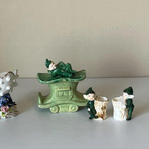 Puede incluir: Colección de figuras de elfos de cerámica. Un elfo descansa sobre una jardinera verde en forma de zapato, mientras que otros están cerca de recipientes con forma de tronco de árbol. Las figuras están pintadas en tonos verdes, blancos y carne, con algunos acentos de rojo y azul.