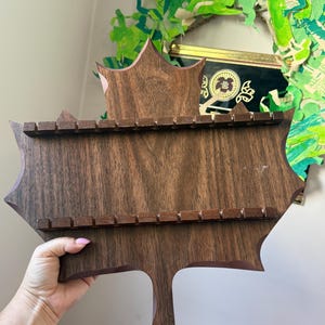 May include: A dark brown wooden display rack shaped like a large maple leaf. The rack features two horizontal bars with evenly spaced notches along the top and bottom sections, designed to hold small items securely in a horizontal arrangement.