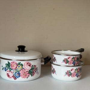 Op de afbeelding: Een witte emaille kookset met een grote pot met deksel en twee kleinere potten. Elk stuk is versierd met een bloemenontwerp in roze, blauw en geel. De set bevat een grote pot met deksel en twee kleinere potten.