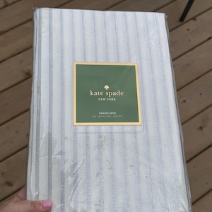 May include: A rectangular tablecloth in a white and grey striped pattern, encased in clear plastic packaging. A green label on the front features the gold text kate spade NEW YORK TABLECLOTH 60 x 84 IN (152 x 213 CM) with a small spade logo.