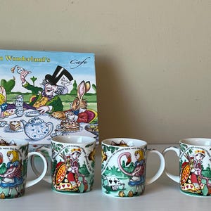 May include: Four colorful ceramic mugs with Alice in Wonderland themed designs. The mugs feature characters like the Queen of Hearts, Alice, and the Mad Hatter. The mugs are all white with green, red, and blue accents. The mugs are arranged in a row on a table.