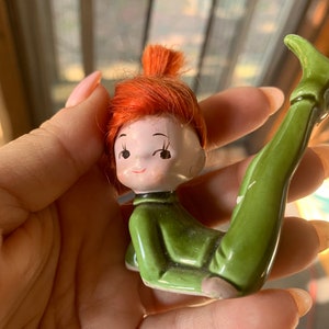 May include: A ceramic figurine of a girl elf with red hair in a ponytail, wearing a green outfit and sitting with her legs stretched out.
