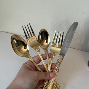 Puede incluir: Un juego de cinco cubiertos dorados, que incluye un cuchillo, dos tenedores y dos cucharas. La cubertería tiene detalles florales ornamentados en los mangos. La hoja del cuchillo es plateada. La cubertería se sostiene en una mano.