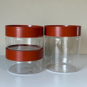 Könnte beinhalten: Drei transparente, zylindrische Aufbewahrungsgläser mit rostfarbenen Deckeln. Zwei Gläser sind gestapelt, das dritte steht daneben. Die Gläser bestehen aus transparentem Kunststoff oder Glas, die Deckel sind einfarbig und undurchsichtig.