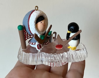 May include: A Christmas ornament featuring a cartoon person wearing a white and grey hooded parka and a penguin playing hockey on a clear ice rink.