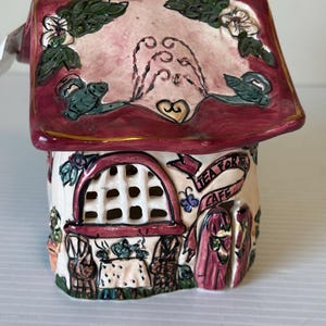 May include: A ceramic house-shaped tea light holder with a red roof and a cream-colored body. The roof is decorated with floral and scroll designs. The body features a window and the text "TEA FOR TWO CAFE" on a banner. The house has a black chimney.