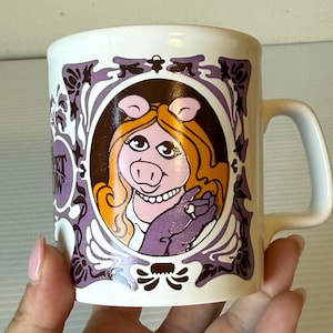 Könnte beinhalten: Weißer Keramikbecher mit einem Cartoon-Bild von Miss Piggy aus der Muppet Show. Das Design umfasst ein Porträt von Miss Piggy in einem lila-braunen Rahmen mit dem Logo der Show. Der Becher hat einen gebogenen Griff.