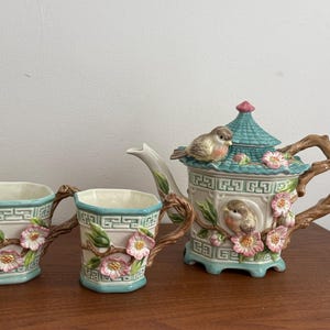 May include: A ceramic teapot and two matching mugs featuring a cream-colored base with a light blue geometric border. The set is decorated with three-dimensional pink flowers, green leaves, and brown tree branch handles. A small bird sits on the lid.