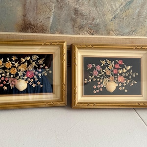 Peut inclure: Deux cadres muraux rectangulaires présentant des compositions florales réalisées à partir de coquillages et de perles sur fond noir. Chaque pièce est encadrée d'une bordure dorée avec un contour intérieur blanc cassé, montrant des fleurs en coquillage.