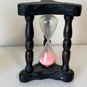 Op de afbeelding: Een klein houten zandloper met een roze zandtimer. De zandloper staat op een zwarte houten standaard met drie poten.