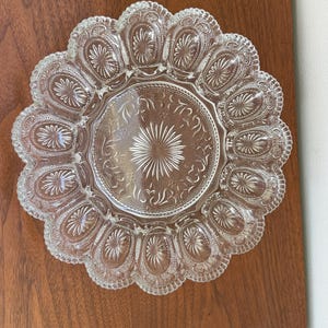 May include: A clear pressed glass serving plate featuring a central starburst pattern surrounded by a ring of twelve oval indentations, each containing a sunburst motif. The scalloped edge is decorated with a fine beaded and textured border.