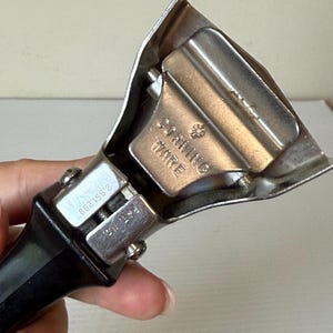 May include: A vintage Corning Ware can opener with a black handle and silver metal components. The can opener has the text "Corning Ware" stamped on the metal. The design includes a cutting wheel and a lever for opening cans.
