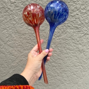 May include: Two hand-blown glass watering globes with long, slender stems. One globe features a vibrant red and orange mottled pattern, while the other displays a deep blue and white marbled design, both ending in a pointed, open-ended tube.