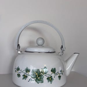 Pode incluir: Bule de chá esmaltado branco com alça e tampa cinzentas. O bule apresenta um design de folhas de hera verdes e amarelas ao redor da base. O bico e a alça são cinzentos, e a tampa tem um pequeno botão redondo.