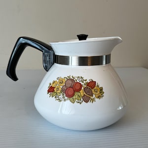 Könnte beinhalten: Weiße Kaffeekanne aus Keramik mit schwarzem Griff und floralem Design mit roten, gelben und braunen Früchten. Die Kanne hat einen silbernen Band um die Oberseite.