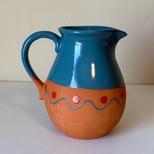 May include: A ceramic jug with a teal blue top and a terracotta-coloured base. The base is decorated with red dots and a wavy blue line. The jug has a curved handle and a spout for pouring.