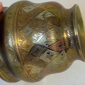 May include: A brass-coloured decorative vase with intricate etched patterns. The vase has a bulbous body and a flared rim. The etched designs include geometric shapes and floral motifs, with hints of pink and silver accents.