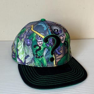 May include: A baseball cap featuring a comic book illustration of the Riddler. The cap has a black brim with green stitching, a green button on top, and a colorful design of the Riddler character on the crown.