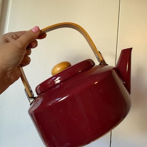 May include: A deep red tea kettle with a glossy finish, featuring a curved light-coloured wooden handle attached with silver metal hardware and a matching round wooden knob on the lid. The kettle has a wide base and a short, angled spout.