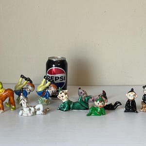 Puede incluir: Una colección de figuras de cerámica vintage, que incluyen acróbatas, elfos y una bruja, en varias poses y atuendos. Se incluye una lata de Pepsi para la escala. Las figuras están pintadas con colores brillantes y detalles.