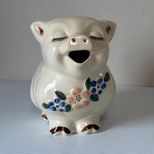 May include: A ceramic pig-shaped pitcher with a cream-colored glaze. The pig has closed eyes, a smiling mouth, and a floral design in blue, pink, and green on its side. The ears and feet are brown.