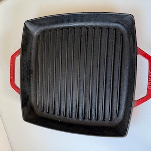 May include: A black cast iron grill pan with red handles. The pan has a square shape with ridged grilling surface. The handles are red and have the word "STAUB" printed on them. This pan is designed for stovetop grilling.