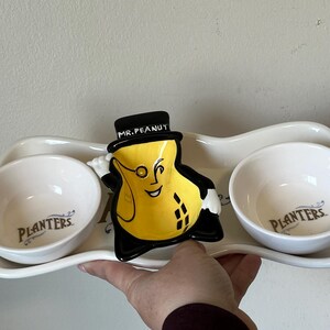 Peut inclure: Un plat de service en céramique avec Mr. Peanut, un personnage de cacahuète jaune avec un chapeau haut de forme et un monocle. Le plat a deux bols blancs avec le logo "Planters" et un design ondulé.