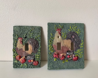 Può includere: Due placche per interruttori con un design a gallo e mele. Il gallo è marrone, bianco e rosso. Le mele sono rosse. Lo sfondo è verde.