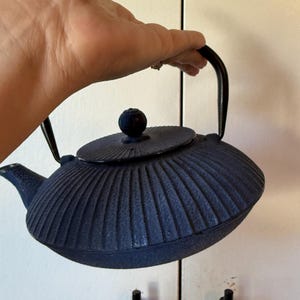 May include: A dark blue cast iron teapot featuring a textured, ribbed exterior surface, a matching circular lid with a small knob handle, and a curved black metal handle attached to the sides of the teapot body for easy lifting and pouring.
