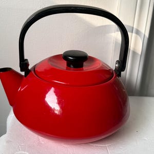 Puede incluir: Una tetera de color rojo brillante con acabado lustroso, que presenta un cuerpo redondeado, un pico corto y un asa arqueada negra. La tapa también es roja con un pequeño pomo negro redondo centrado en la parte superior del recipiente.