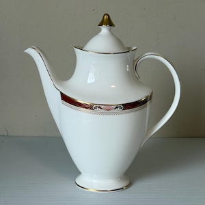 May include: White porcelain coffee pot with a gold-colored finial and trim. The pot features a decorative band of red, gold, and green floral designs. The handle and spout are curved, and the pot sits on a small foot.