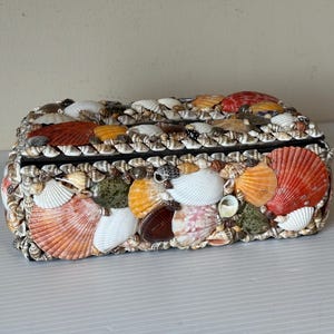 May include: Rectangular box covered in various seashells. The box features a black border and is adorned with a variety of colorful shells, including orange, white, and red. The shells are arranged to create a textured surface.