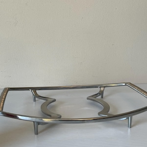 Puede incluir: Un soporte de bandeja de servicio de metal plateado. El soporte tiene un marco rectangular curvo con asas decorativas y dos soportes curvos. Tiene cuatro patas cortas para mayor estabilidad. El soporte está diseñado para sostener una fuente.
