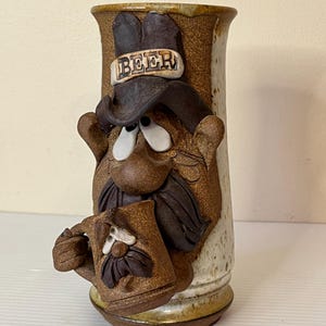 May include: A ceramic beer mug with a cartoon face, a brown hat with the word "BEER" on it, and a mustache. The mug is holding a smaller mug in its hand.