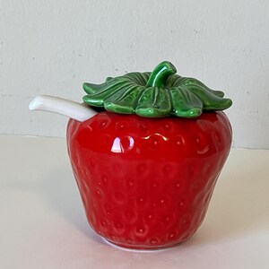 May include: A red ceramic strawberry-shaped sugar dispenser with a green leaf lid and a white spout.