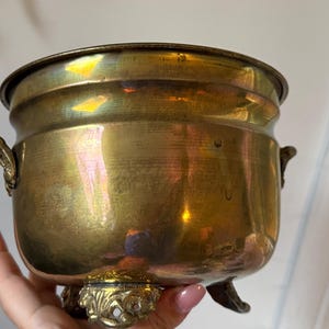 May include: A round brass pot with a polished metallic finish, featuring two small decorative side handles and three ornate, sculpted feet. The vessel has a wide, open top and a smooth, curved body that reflects light across its surface.